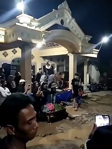 A nighttime scene unfolds, capturing a live musical performance in front of a house. A woman in a dark top stands on an elevated porch or balcony, singing into a microphone, with speakers visible behind her. This elevated area functions as a makeshift stage. Below and around the stage, a crowd of people gathers, watching the performance. Some individuals in the foreground are holding up their phones, seemingly recording the event. Two women in the middle foreground wear matching black t-shirts, one with text that appears to read "SENTRI" and more beneath it, and both are attentively watching the singer. A man in a black t-shirt and shorts stands to the right. Artificial lights illuminate the entire scene, confirming it is dark outside. Other musical equipment, including what looks like a keyboard on a table covered with a blue tarp, can be seen near the stage. The overall atmosphere suggests a community gathering or local event.