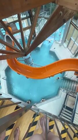 From a high, angled perspective, a child with arm floaties slides down an orange water slide into a pool. The pool is surrounded by people swimming and relaxing. The camera is positioned on a staircase with black and yellow caution tape, and portions of a wood structure are visible in the frame. The pool area is bright and modern, with natural light streaming through large windows. The water in the pool appears to be a clear, light blue.