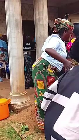 The video captures a group of women gathered outside a building, possibly at an event or celebration. One woman, dressed in a green and yellow patterned skirt and a light blue top, is at the center of attention. Other women are nearby, some in brightly colored outfits. One woman holds up a smartphone, possibly filming or taking pictures. The environment includes the exterior of a building with pillars and some visible equipment labeled "DJ LASKI". The atmosphere seems lively, and there is likely music playing in the background.
