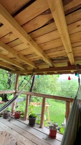 The video shows a view from under a wooden porch roof, where someone is relaxing in a hammock. The individual is wearing black socks and their legs are visible. The porch has a wooden railing with potted plants lining it. String lights hang below the roof. Beyond the railing is a green, grassy lawn and trees. In the distance, there are mountains. The camera angle is low, looking up and out from inside the porch.