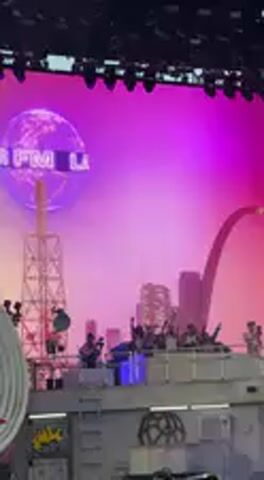 This video captures a vibrant night scene at what appears to be a large music festival or concert. The primary focus is on a substantial crowd of attendees, many of whom are holding up their smartphones to record the performance.

In the background, a large stage features a prominent, multi-level, grey industrial-style structure, possibly resembling a tank or a futuristic building. Several performers or crew members can be seen on top of this structure. Behind the stage is a massive video screen or backdrop illuminated with dynamic purple and pink lighting. This screen displays a stylized cityscape silhouette, notably featuring a large archway that resembles the Gateway Arch in St. Louis, Missouri, and a tower akin to the Eiffel Tower. A glowing purple sphere with partially visible text, possibly "LAX," is also displayed on the screen, matching text visible on the lower left of the physical stage structure.

The atmosphere is energetic, with the crowd engaged in the event. The lighting suggests it is either an evening outdoor event or an indoor venue designed to replicate a night setting. The location context is Indio, United States, likely referring to the Coachella music festival, and the stage design elements are part of the artistic production.