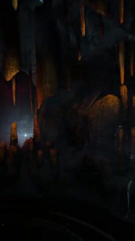 The video shows a dark, cavernous environment illuminated by various colored lights, primarily orange and red, casting shadows on stalactite and stalagmite formations. The camera appears to be moving, possibly on a ride or vehicle, as evidenced by the blurred motion and the shifting perspective. In the background, faint shapes suggest the presence of figures or props, some appearing as silhouettes. The overall atmosphere is dim and mysterious, with the lighting creating an otherworldly effect on the rock structures.