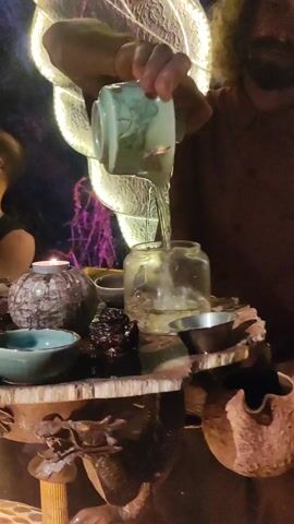 A man pours liquid from a small green ceramic teapot into a glass jar on a wood table. The man has dark curly hair and a beard and is wearing a brown shirt. The table is decorated with several small bowls, a rock, and a lit candle. A decorative light fixture is in the background. The background is dimly lit and appears to be indoors. The man’s movements are slow and deliberate.