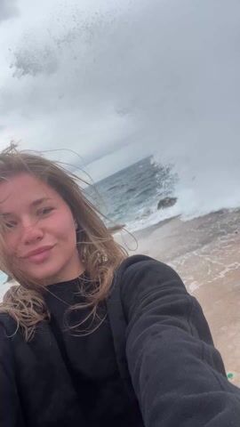 The video shows a fair-skinned woman with shoulder-length blonde hair standing on a beach. She is wearing a black sweatshirt. The wind is blowing her hair across her face. Behind her, waves are crashing against rocks on the shore. The sky is overcast, and mountains are visible in the distance. She looks directly at the camera, speaks briefly, and then looks down. The camera appears to be hand-held, as it shifts and moves slightly throughout the video.