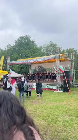 A medium shot shows a group of Bulgarian folk singers, with one instrument player, performing on an outdoor stage. They are singing in unison, dressed in traditional clothing, primarily black, white, and red. A backdrop behind them shows a ram's head. Banners in Bulgarian hang above the stage. In front of the stage, a small crowd watches. Some people are dressed in rain gear, indicating wet weather. A Bulgarian flag is visible to the right of the stage. The stage is set up on a grassy field. The sky is overcast.