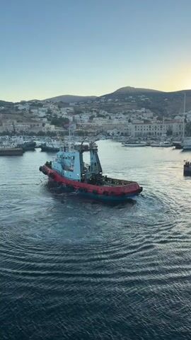 The video shows a tugboat in a harbor. The tugboat is red and blue and is turning in a circle, creating ripples in the water. In the background, there is a town with white buildings and a hill. The sky is blue and clear.