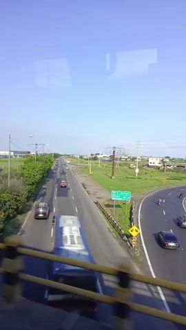 The video shows a highway from an elevated position. The road is straight and has multiple lanes, with cars and trucks driving on it. On either side of the highway is grassy land with scattered trees and some buildings in the distance. The sky is clear and blue. The view is partially obstructed by a yellow railing in the foreground and reflections on the window the video is taken through. The camera appears to be moving, likely from a vehicle passing over a bridge.