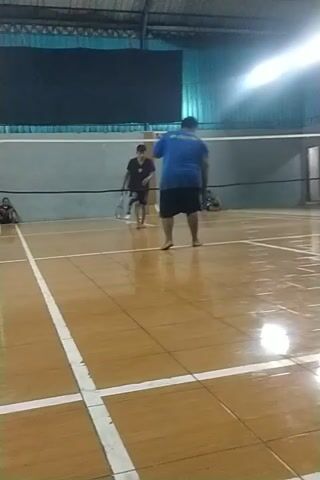 The video captures an indoor badminton court with a wooden floor. Two individuals are present. One person, wearing a blue t-shirt and black shorts, is standing near the net. Another person, in a white t-shirt and black shorts, is positioned further back on the court. The lighting in the court is bright, with lights visible on the ceiling. The court markings, including white lines, are clearly visible. The scene appears to be a pause or preparation for a badminton game.