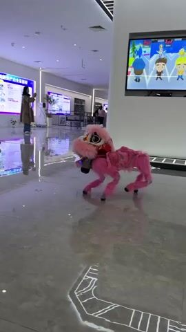 A small, pink, four-legged robotic animal, resembling a Chinese dragon costume, performs a handstand in a modern, brightly lit indoor space with a polished tile floor. A woman in a tan coat and jeans looks at her phone while standing in the background. A child in a blue vest walks into the frame from the left. A large television screen on the wall displays a colorful, cartoon-like game. Signs with Chinese characters are visible in the background. The floor reflects the lights and the robot.
