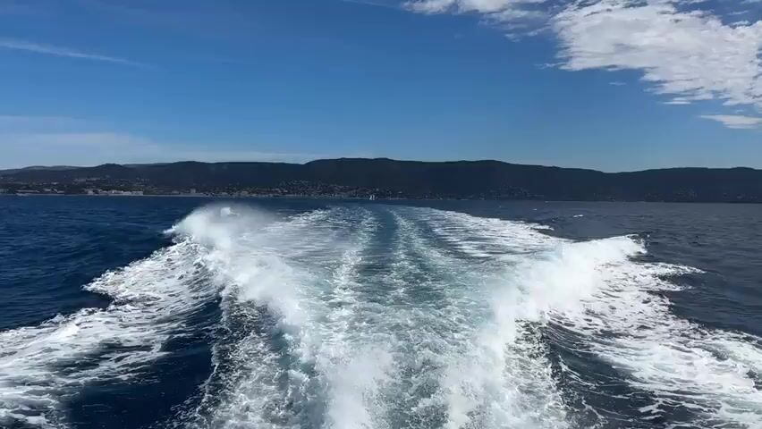 The video captures the wake of a boat moving through the ocean under a clear blue sky. The camera is positioned looking backward from the boat, focusing on the turbulent water churned up by its passage. White foam and waves stretch out behind the vessel, creating a distinct V-shaped pattern. In the distance, a hazy coastline with hills and buildings is visible, separated from the water by a strip of land. The sea is a deep blue, contrasting with the bright white of the wake. The overall impression is one of forward motion and open water.