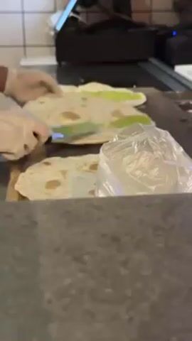 A close up shot shows a worker’s hands wearing white gloves preparing flatbreads. The flatbreads are arranged in a row on a dark surface. Two of the flatbreads have a green substance spread on them. Next to the last flatbread is a plastic bag. The worker spreads the green substance on the last flatbread. In the background is a tile wall, a point of sale system, and other kitchen equipment.