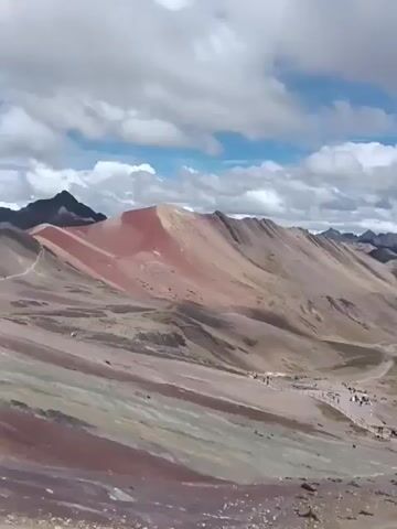 The video shows a mountainous landscape under a partly cloudy sky. In the foreground, a trail with a wooden barrier leads towards distant mountains. Numerous people are walking along the trail. The mountains exhibit a variety of colors, including red, brown, and grey, with some snow visible on the peaks of the more distant mountains. The clouds are white and fluffy, with patches of blue sky visible.