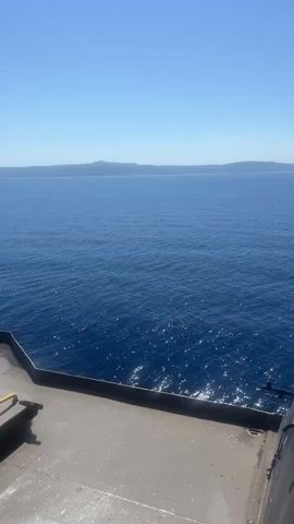 The video shows a wide expanse of deep blue ocean under a clear, bright blue sky. The sun glints off the water's surface, creating a shimmering effect. In the foreground, a gray deck of a ship is visible, with a dark-colored structure and a metal hatch with yellow handles. A thick rope extends vertically from the top of the frame downwards, casting a shadow. In the distance, a faint, hazy line of land is visible on the horizon. The scene suggests a calm day at sea.