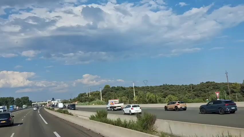 The video captures a view from a vehicle traveling on a highway. The highway appears to be a multi-lane road with a concrete barrier separating traffic in opposite directions. Several vehicles are visible, including cars, trucks, and what appears to be a small utility truck or van. On the right side of the highway, there is a dense line of green trees and vegetation, suggesting a rural or semi-rural environment. The sky is blue with scattered white clouds, indicating fair weather. In the distance, there's a slight incline of land covered in trees. There are also utility poles visible along the side of the road. The footage shows traffic moving along the highway, with vehicles in different lanes and at varying distances. The perspective is from the driver's seat or a passenger in the front seat, looking forward.