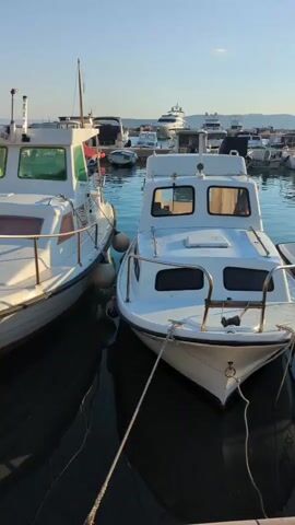 The video shows a marina with several boats docked. The foreground features two white motorboats tied together. The boat on the left is closer to the camera and is partially visible. The boat on the right is also white and has the registration number MA 249 visible. Another smaller white boat with the registration number MA 1004 is nestled next to it. In the background, a variety of other boats, including sailboats and larger yachts, are moored. The sky is clear with some clouds, and the sun appears to be setting, casting a warm light on the scene. The water is calm and reflects the boats and the sky.