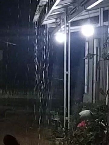 The video shows a dark, rainy night. Water streams down from an overhang, likely a roof or shelter. Two lights are visible, illuminating the area with a bright, white glow. In the foreground, a dark, rounded object, possibly a motorcycle seat, is visible. Beyond the overhang, a building with vertical blinds is seen, with lights on inside. The ground appears wet and reflects the light. There are plants and foliage around the building. The overall impression is of a dimly lit, outdoor scene during a downpour.
