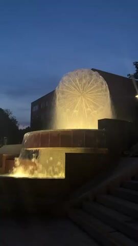 A fountain with a spherical, dandelion-like water feature is illuminated with yellow lights. Water cascades down a tiered structure next to it, and stairs are visible on the right. The scene is set at dusk or night, with a dark blue sky and the lights of a building in the background.