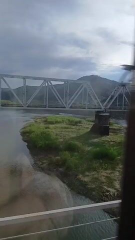 From the window of a train, a metal truss bridge is visible, spanning a river or wide stream. The bank is sandy and covered in green grass. A low mountain is in the background, under an overcast sky. The camera moves as the train passes.