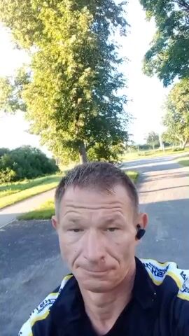 A close-up shot shows a man with short, blondish-brown hair, a fair complexion, and weathered skin. He is wearing a black jacket with yellow and white detailing. He has earbuds in both ears. The man is looking directly at the camera. In the background, a paved path curves into the distance, lined with green grass and trees. The sky is bright and appears to be daytime. A large, silver, curved building with a door is visible on the left side of the frame. The man's head and shoulders fill most of the frame.