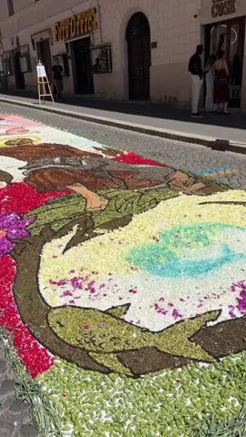 A street view shows a long artwork made of flower petals spread across a cobblestone street. The artwork depicts a figure with a halo, a wolf, and floral designs in various colors. A man in a black T-shirt and light-colored pants stands beside an easel on the sidewalk to the left, observing the artwork. Another man in a black shirt and a woman in a light-colored dress are seen on the right sidewalk. The buildings lining the street have shop signs, and the sun casts shadows, indicating a bright day.