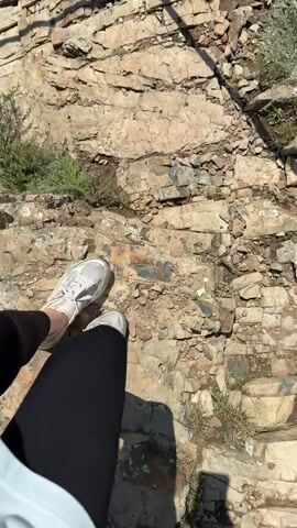 The video shows a first-person perspective of someone sitting on a ledge of rocks overlooking a rocky terrain. The person is wearing black leggings and white sneakers. Sunlight casts shadows on the ground. The camera then pans down, showing the person standing up and stepping onto a concrete path with cracks. The path appears to be outdoors, with rocks and vegetation surrounding it. The video is short and focuses on the act of standing up and moving onto the path.
