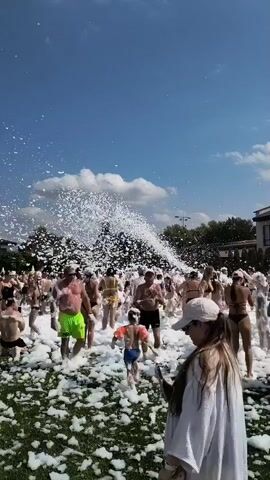 A crowded outdoor foam party is in full swing under a bright blue sky with scattered clouds. A powerful foam machine sprays a massive cloud of white foam into the air from the left side of the frame, covering the ground and most of the people. Many individuals, mostly young adults, are dressed in swimwear and are actively participating in the foam, with some dancing and playing. In the foreground, a woman wearing a white hat and a white cover-up is looking at her phone, partially obscuring the view of the party. The ground appears to be covered in artificial turf, and in the background, trees and a building are visible. The overall atmosphere seems to be one of energetic revelry and fun.