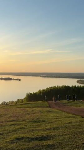 The video shows a panoramic view of a wide river at sunset. The sky transitions from blue to orange and yellow, reflecting in the calm water. Several islands dot the river's surface. A grassy hill with a dirt path in the foreground is populated with people, some standing near the edge, enjoying the view. The scene is serene and peaceful.