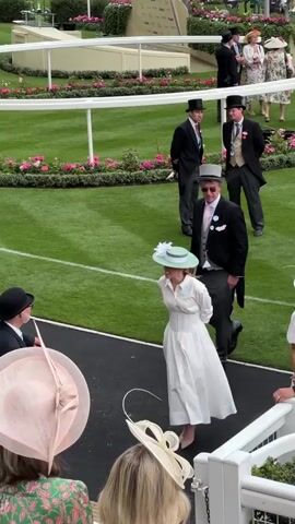This video captures a moment at a formal outdoor event, likely a racecourse given the manicured grass and white fencing in the background. Several individuals are dressed in formal attire. In the foreground, a woman in a light blue dress and a wide-brimmed hat stands to the right, facing left. To her left, a man in a black tuxedo with a gray top hat and a pink tie walks away from the camera. A woman in a white dress and a mint green hat is walking towards the man. To the left of the woman in the white dress, a man in a black bowler hat and a suit faces the man in the tuxedo. In the middle ground, two men in similar formal attire, including top hats and waistcoats, stand on the grass. Further back, two more men in formal wear are visible. The overall atmosphere is one of a sophisticated gathering.