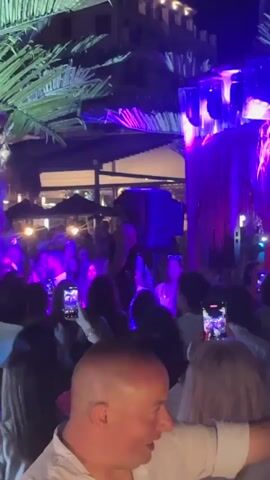 The video shows a crowded outdoor event at night, illuminated by colorful stage lights. A male performer, wearing a black tank top and sunglasses, is on a raised platform, holding a microphone and gesturing to the audience. The audience members, facing the performer, are holding up their phones to record. Palm trees are visible in the background, suggesting a tropical or beachside location. The lighting shifts between purple, red, green, and blue, creating a dynamic atmosphere. In the foreground, several audience members are visible, including a man in a white shirt and a woman with blonde hair. The overall scene depicts a live music performance or DJ set in a vibrant, energetic setting.