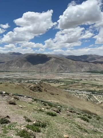 The camera, positioned on a grassy slope, offers a wide-angle view of a valley and mountain range under a partly cloudy sky. The valley contains fields, a road, a river, and some structures. In the foreground, sparse vegetation dots the hillside. The mountains in the background are a muted brown and green. The sky is a bright blue, with fluffy white clouds scattered across it. The sunlight casts shadows on the landscape, creating a contrast between light and dark areas.