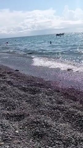 The video shows a rocky beach with people swimming in the ocean. There are waves gently rolling onto the shore. In the distance, a small boat is visible on the water. The sky is blue with some white clouds. The water appears clear and blue.