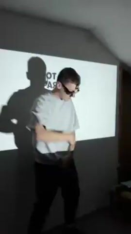A young man dances in front of a wall with words projected on it. He is wearing a gray shirt covered with black text, black pants, and dark glasses. His shadow appears on the wall. He moves his hands and body to the beat of a song. The lighting changes in the room as the video continues. The background features a partially visible wooden structure.