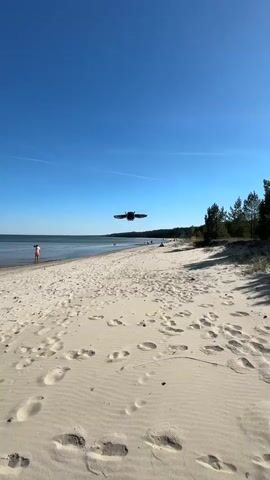 The video shows a view of a sandy beach under a clear blue sky. A drone hovers in the air above the beach.  A person's hand is visible in the foreground, briefly obscuring the view before being lowered. In the background, people are walking on the beach and standing near the water.  A line of trees borders the beach on one side. Footprints are visible in the sand.  The sun shines brightly.