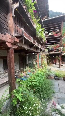 The video shows a rustic, multi-story wooden building with balconies overflowing with potted plants and flowers. In the foreground, a cobblestone path winds through a lush garden filled with various plants and flowers in pots. Behind the wooden building, there are stone structures, including old fountains or troughs, and a steep, tree-covered mountain rises in the background under a cloudy sky. The scene appears to be a quaint, historical village or farmstead, likely in a mountainous region. There are no people visible in the video. The camera pans slowly across the scene, providing a detailed view of the architecture, vegetation, and surrounding landscape.