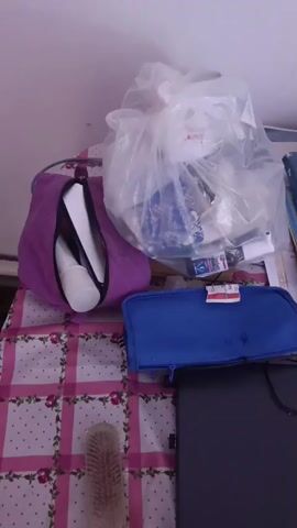 The video shows a cluttered desk surface covered with a pink and white checkered tablecloth. On the desk are various items including a purple cosmetic bag with toiletries peeking out, a clear plastic bag filled with assorted items, a blue pencil case, a black laptop, a hairbrush, a stack of books with a pair of glasses on top, a green bottle, and a computer mouse. The camera pans across the desk, showing the items from different angles. The lighting suggests an indoor environment.