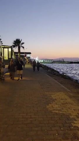 The video shows a waterfront promenade at dusk. On the left, a paved walkway is lined with people strolling. A palm tree stands tall near the beginning of the path. Further along the walkway, buildings are visible. The right side of the video is dominated by the dark blue sea, with gentle waves lapping against a rocky shoreline. The sky above is a soft gradient of twilight colors, transitioning from pale orange near the horizon to a deep blue. A distant structure, possibly a lighthouse, can be seen on the water in the far distance. The overall atmosphere is peaceful and serene, with the warm glow of artificial lights illuminating the walkway.