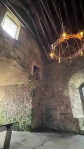 The video shows the interior of a stone structure, possibly a castle or old building. The room has stone walls with arched windows and a wooden-beamed ceiling. Two decorative, circular chandeliers with what appear to be electric candles hang from the ceiling. A large wooden table sits in the center of the room. The stone floor is gray and appears to be uneven. The lighting is dim, primarily from the windows and chandeliers. The camera slowly pans across the room, showcasing the architectural details and the age of the space.