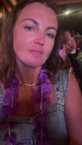 The video shows a close-up of a woman with brown hair, wearing a denim overall dress and a purple lei. She is in what appears to be an outdoor setting with other people visible in the background. The lighting suggests it is nighttime, with some artificial lights visible in the upper background. The woman is speaking and gesturing, with her mouth open in some frames, indicating she is actively engaged in conversation or presenting something.

The scene seems to be a social gathering or event, possibly a luau or a celebration, given the lei. The woman's expressions shift from smiling and blowing a kiss to speaking animatedly, suggesting a lively and engaging atmosphere. The background is blurred but shows other patrons seated at tables, further supporting the idea of a restaurant, bar, or event venue. The woman's attire and the lei contribute to a festive, possibly tropical, ambiance. The overall mood appears to be upbeat and enjoyable. There is no visible text in the media. The context provided places this scene in La Grande-Motte, France.