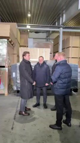Four men are standing in what appears to be a warehouse or storage facility. One man in a long black coat shakes hands with a man in a shorter black puffer jacket. They turn to face a large, machine-like object wrapped in plastic, which is in front of a wooden crate. Two other men stand nearby, also wearing dark jackets. The background shows stacks of packaged materials. The lighting is bright, suggesting an indoor setting.