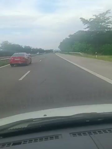 This video shows a view from inside a car driving on a highway. The road is paved with clear white lane markings, and there are other vehicles on the highway, including a red car ahead of the viewer's car and some white vans further in the distance. The scene is flanked by a grassy shoulder and a tree line on the right side of the road, with more trees and foliage visible on the left. The sky is overcast, suggesting a cloudy or partially cloudy day. The dashboard of the car is visible in the foreground, indicating the perspective is from the driver's or passenger's seat. The overall mood is one of travel and movement. There is no visible text in the video. The location context provided is Gorno Varshilo, Bulgaria.