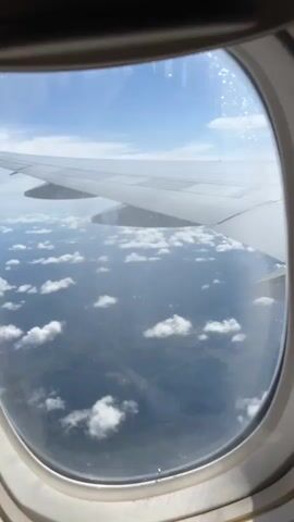 A view from an airplane window shows a landscape covered in scattered clouds and a body of water. An airplane wing is visible in the upper right corner of the frame. The sky is blue with some wispy white clouds. The footage appears to be taken during the daytime.