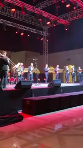 A big band with approximately ten members, all dressed in suits, is performing on a brightly lit outdoor stage at night. The stage has red lighting, and there is a large speaker on the left-hand side. The band includes wind instruments, and drums. The band appears to be playing in a plaza or outdoor space with building visible in the background.