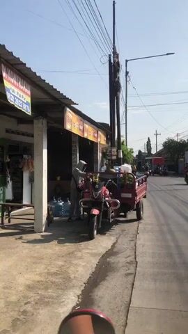 The video shows a street scene. A red motorcycle with a trailer is parked on the side of the road. The trailer is loaded with several large water jugs and gas cylinders. A person wearing a grey hooded jacket and a blue bandana over their face is standing next to the motorcycle, appearing to be loading or unloading items from the trailer. A small shop with a yellow awning is visible behind them, with signs advertising products. The street is paved, and there is a sidewalk. In the background, other vehicles and people can be seen on the street. Power lines and utility poles are visible overhead. The sky is clear and blue, indicating daylight.