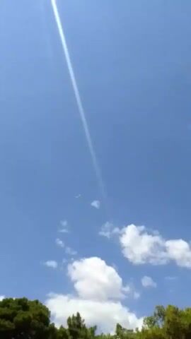 The video shows a bright blue sky with a few scattered white clouds. A beam of sunlight cuts diagonally across the frame from the top left. In the distance, a small, white object, likely an airplane, can be seen flying. The bottom of the frame shows the tops of green trees.