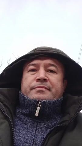 A close-up shot shows a man from the chest up, wearing a dark green hooded jacket and a blue patterned turtleneck sweater. The hood is up, framing his face. He looks directly at the camera with a neutral expression. The background is a blurred, overcast sky. Sounds of children are audible.