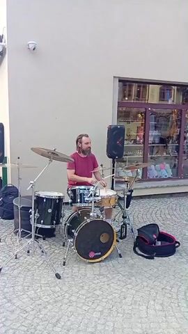 A male drummer with a beard and dreadlocks plays a drum set on a cobblestone street in front of a building with large windows. He is wearing a maroon t-shirt and shorts and is seated behind the drums. A black backpack rests on the ground to his left, and another bag, red and black, is to his right. Two black speakers are positioned on either side of the drumming area. A person in a black top and jeans walks past the drummer from right to left. The scene appears to be an outdoor performance.