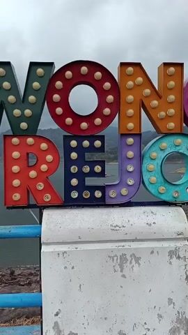 A vertical video shows a sign with large, colorful, marquee-style letters that spell out "WONOREJO". The letters are lit with small, round bulbs of various colors. The sign is mounted on a white, weathered concrete base. Behind the sign, a body of water, likely a lake or reservoir, is visible, with hills or mountains in the distance under a cloudy sky. A blue and white railing runs along the edge of what appears to be a walkway or bridge, in front of the sign and the water. The camera pans slightly to the right, showing more of the railing and the water, with the sign remaining in the frame. No people are clearly visible in the video.