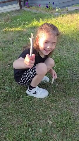 A young girl with long, curly brown hair and a bright smile is playing outside in a grassy yard. She is wearing a black t-shirt and black and white patterned shorts, along with white sneakers and black socks. In the video, she is seen squatting down, holding two sticks. She waves at the camera and then throws one of the sticks, before standing up and turning away. The background shows a wooden fence, a building, and some flowers. The lighting suggests it is daytime, possibly late afternoon or early evening.