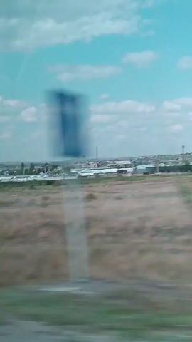 The video shows a landscape with a cloudy sky. In the foreground, there is dry grass and some green grass. In the middle ground, there are some buildings and structures that appear to be industrial or agricultural. There are also trees and bushes visible. The background shows more buildings and structures stretching into the distance. The video appears to be filmed from a moving vehicle, as evidenced by the blurriness and the sense of motion. The overall impression is of a rural or semi-industrial area on a clear day.