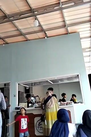 A group of people are inside a brightly lit indoor space, likely a cafe or food establishment, with a high ceiling constructed of metal beams and translucent panels allowing natural light to filter through. In the center of the frame, a light blue wall serves as a backdrop for a counter area where two employees wearing dark shirts and caps are visible. One employee is behind the counter, seemingly preparing food or drinks, while another stands to the right, attending to a customer. Several customers are also present. To the left, a young girl in a red t-shirt stands near a refrigerator displaying various food items. A woman in a grey hijab stands in front of the refrigerator. Further to the right, a woman in a beige and yellow patterned abaya, also wearing a hijab, is interacting with an employee at the counter. Another person, obscured by the foreground, is also present. A price list for frozen food is displayed on a red and white sign. The overall atmosphere appears casual and busy.